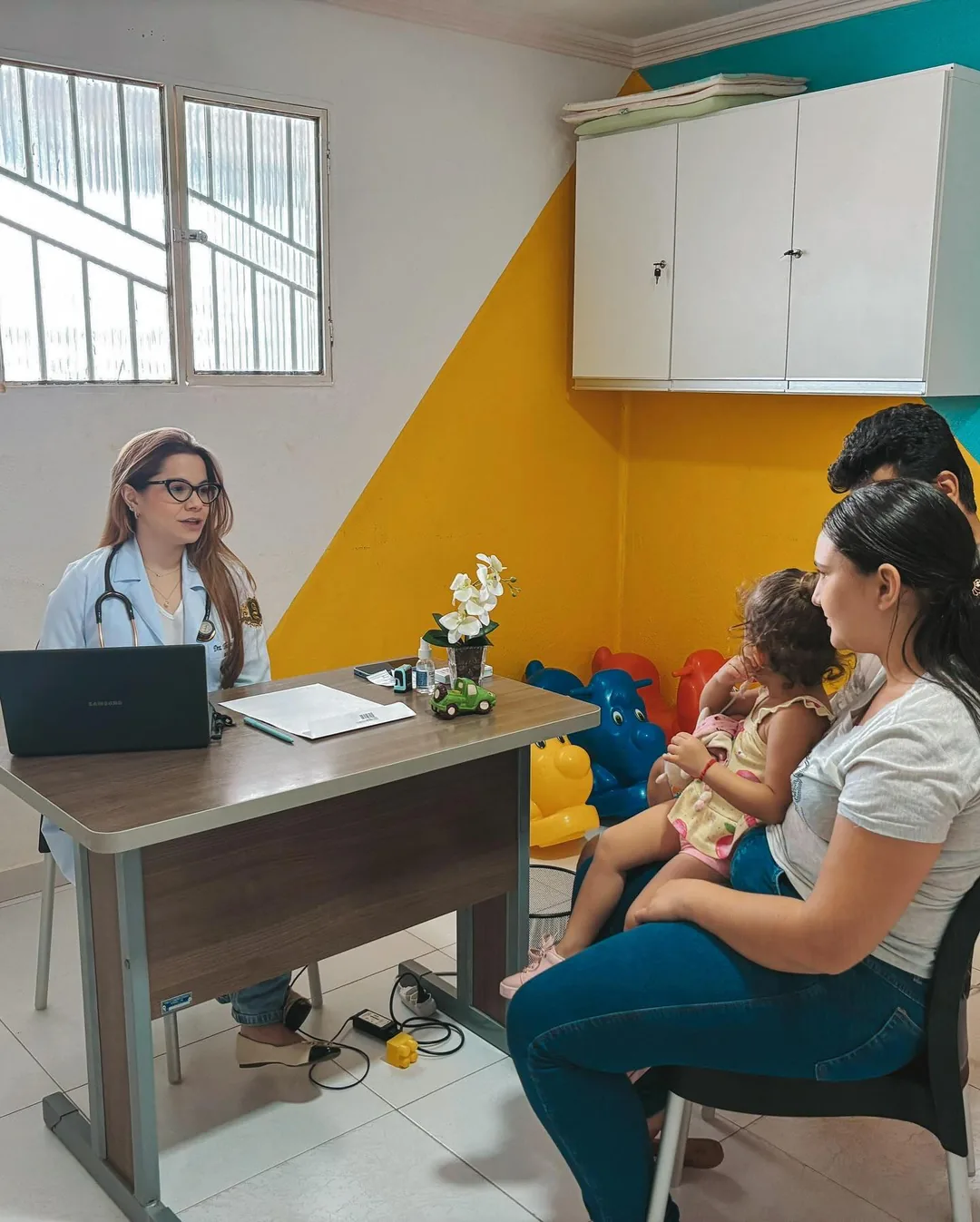 Médica do projeto O Bom Samaritano atende uma família, com a criança no colo da mãe, em sala colorida