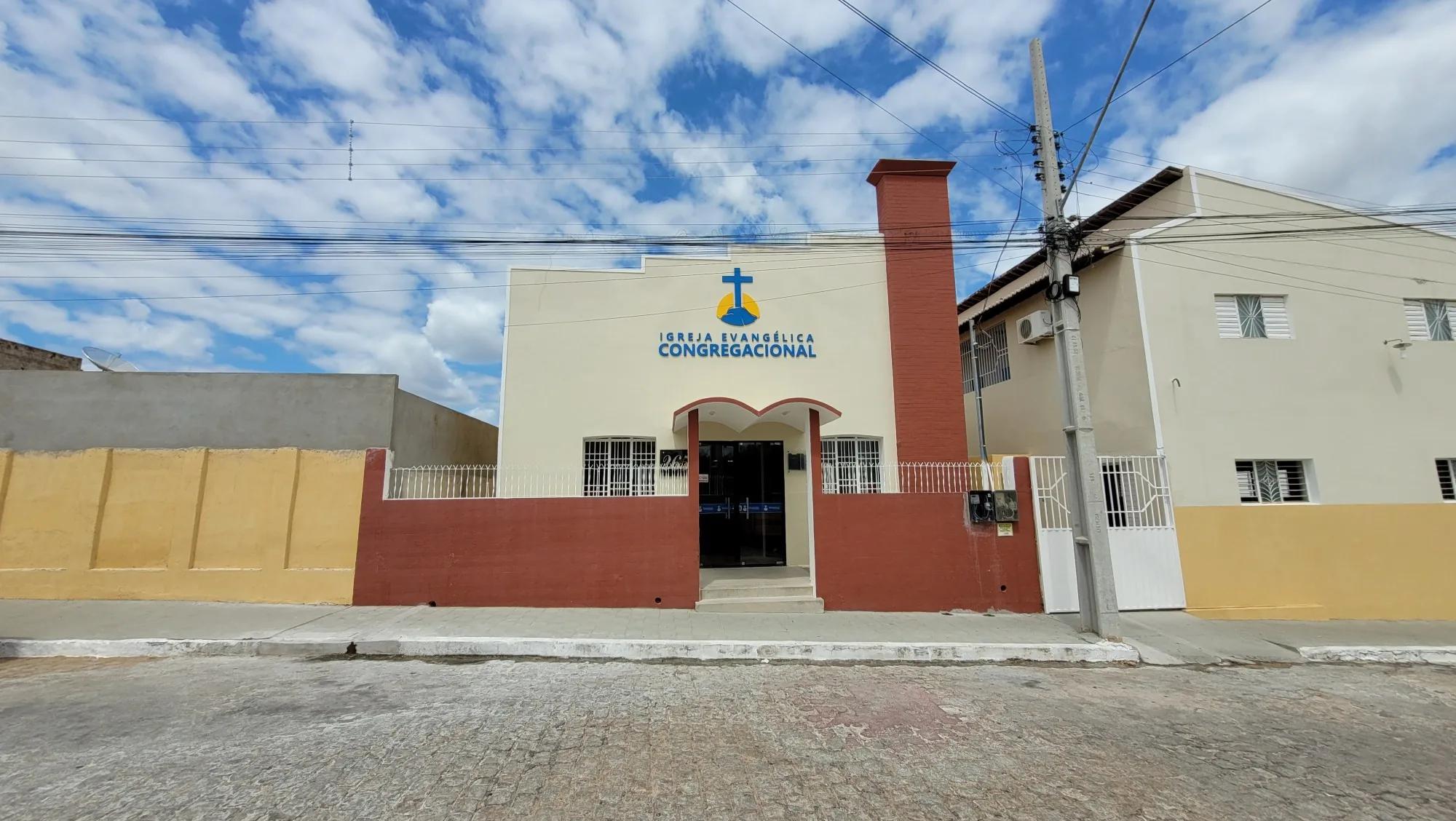Vista da rua com a fachada do templo da Igreja Evangélica Congregacional de Pombal, de muros vermelhos e paredes creme