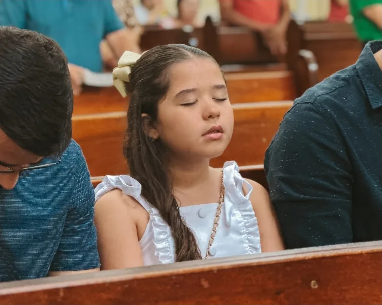 Menina de olhos fechados em oração, sentada entre adultos no banco da igreja
